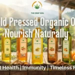 Five bottles of The Deep Soil cold-pressed organic oils—hemp, peanut, mustard, almond, and apricot—lined up on a wooden table against a sunny mountain landscape.
